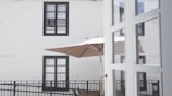 A view of a simple outdoor space with a large beige patio umbrella. The umbrella is set up on a balcony-like area with a black railing. Two rectangular windows with black frames are visible on the light-colored wall behind the balcony. Part of a white door with glass panels is visible in the foreground.