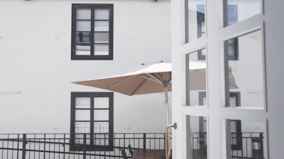 A view of a simple outdoor space with a large beige patio umbrella. The umbrella is set up on a balcony-like area with a black railing. Two rectangular windows with black frames are visible on the light-colored wall behind the balcony. Part of a white door with glass panels is visible in the foreground.