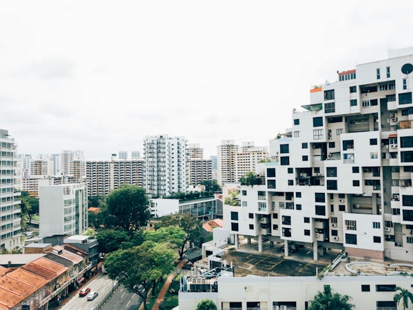 A cityscape with modern, uniquely structured apartment buildings surrounded by high-rise residential towers. Streets with vehicles and rows of trees add a touch of greenery. The architecture features a blend of geometric patterns, and the scene exudes a mix of urban and natural elements.