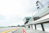 A modern racing track with a building featuring large, closed garage doors. In the background, there is a Ferris wheel towering over the scene along with unique architectural structures resembling domes. The skies are mostly cloudy with a hint of sunlight.