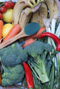 A variety of fresh produce including bananas, lemons, tomatoes, carrots, broccoli, red chili peppers, green onions, and a wooden spoon with a green powder.