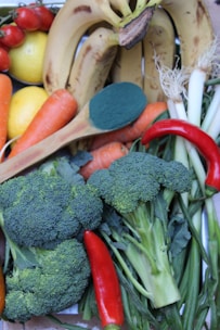 A variety of fresh produce including bananas, lemons, tomatoes, carrots, broccoli, red chili peppers, green onions, and a wooden spoon with a green powder.