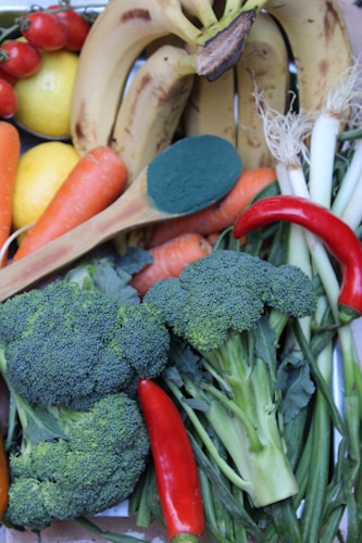A variety of fresh produce including bananas, lemons, tomatoes, carrots, broccoli, red chili peppers, green onions, and a wooden spoon with a green powder.
