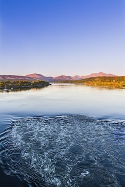 A gentle sky blue river winding toward the viewer with misty gray mountains and a bright yellow sun rising behind them.