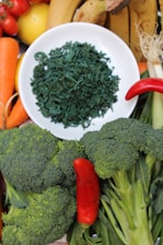 A variety of fresh fruits and vegetables are arranged around a white plate filled with green dried spirulina. The assortment includes bananas, carrots, cherry tomatoes, a lemon, a red chili pepper, and broccoli.