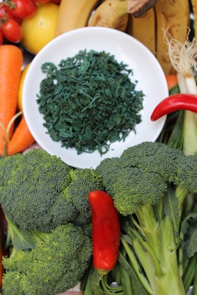 A variety of fresh fruits and vegetables are arranged around a white plate filled with green dried spirulina. The assortment includes bananas, carrots, cherry tomatoes, a lemon, a red chili pepper, and broccoli.