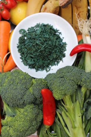 A variety of fresh fruits and vegetables are arranged around a white plate filled with green dried spirulina. The assortment includes bananas, carrots, cherry tomatoes, a lemon, a red chili pepper, and broccoli.