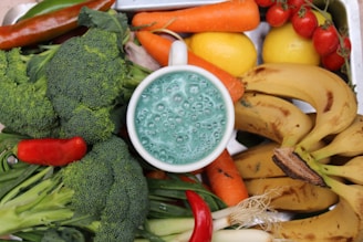 A collection of fresh fruits and vegetables surrounds a cup filled with a foamy, blue-green liquid. The produce includes broccoli, carrots, bananas, lemons, red chili peppers, green onions, and cherry tomatoes.