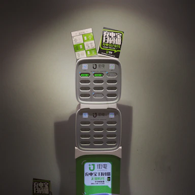 A charging station with multiple slots for charging devices, featuring two advertising flyers attached to the top. The station displays a logo and has various buttons and indicators.
