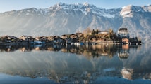 A serene lakeside village is nestled at the foot of majestic snow-capped mountains. The calm water reflects the charming houses and an elegant, castle-like building. The scene is bathed in soft sunlight, enhancing the tranquil atmosphere.