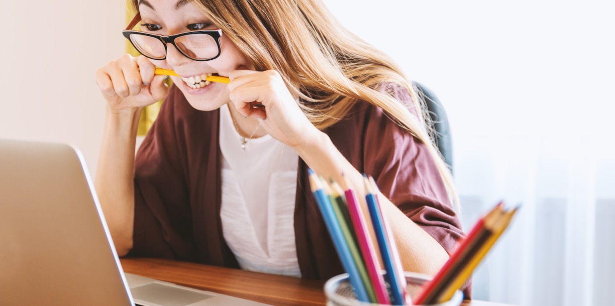 woman biting pencil while sitting on chair in front of computer during daytime