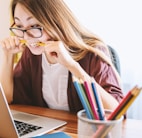 woman biting pencil while sitting on chair in front of computer during daytime