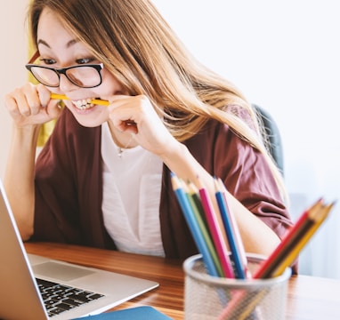 woman biting pencil while sitting on chair in front of computer during daytime