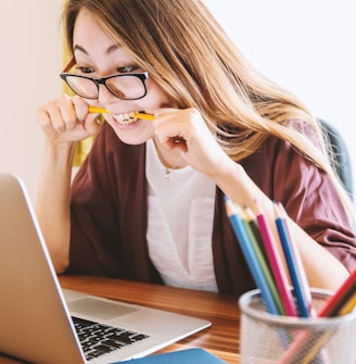 woman biting pencil while sitting on chair in front of computer during daytime