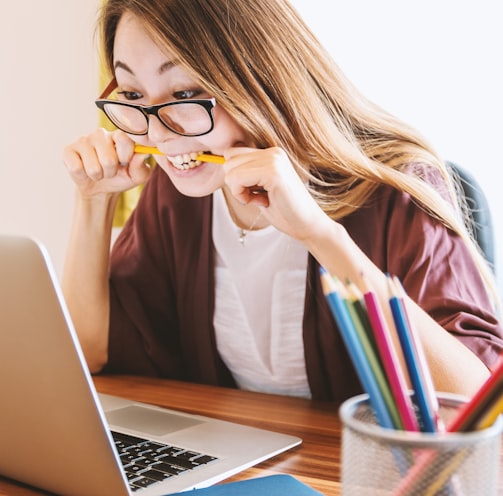 woman biting pencil while sitting on chair in front of computer during daytime