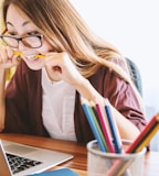 woman biting pencil while sitting on chair in front of computer during daytime