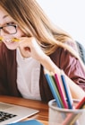 woman biting pencil while sitting on chair in front of computer during daytime