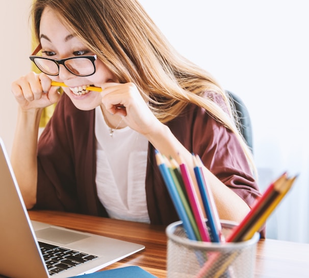 woman biting pencil while sitting on chair in front of computer during daytime