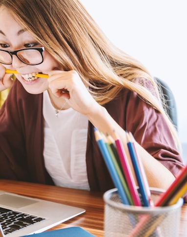 woman biting pencil while sitting on chair in front of computer during daytime