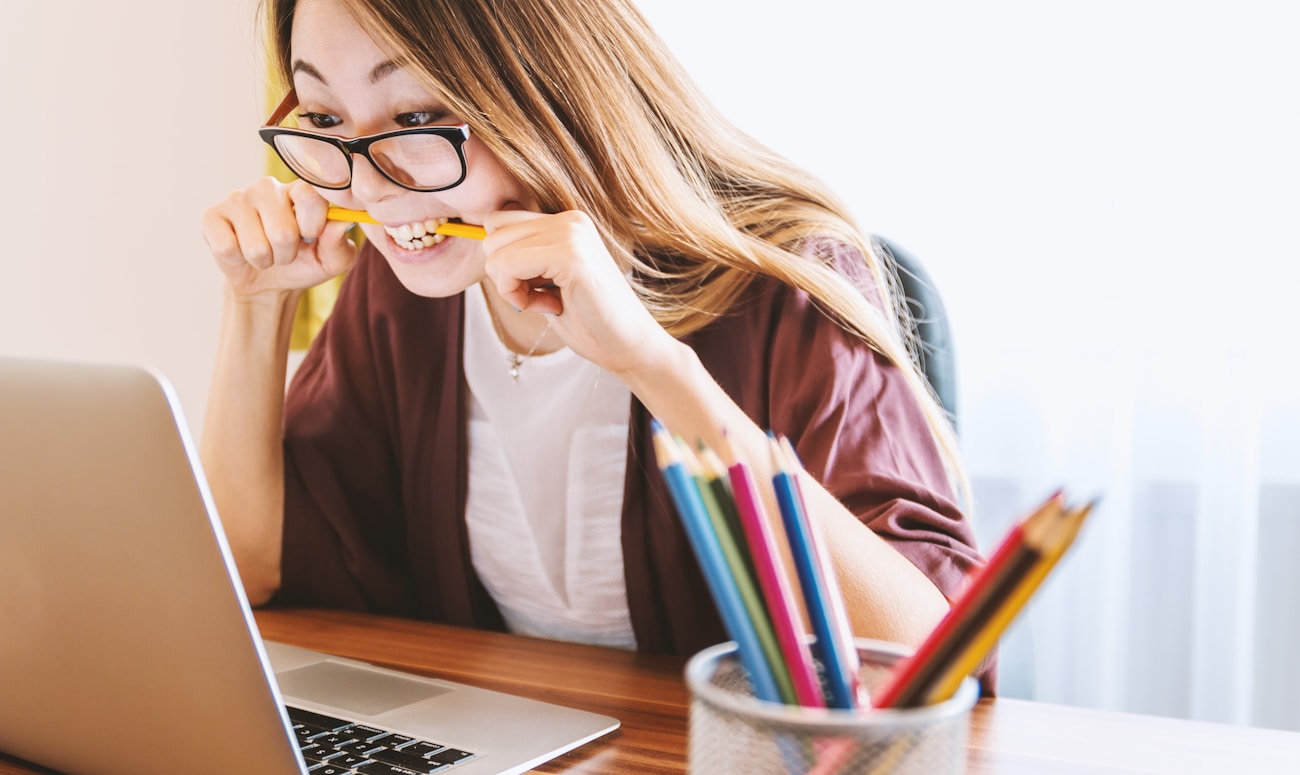 woman biting pencil while sitting on chair in front of computer during daytime