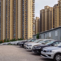 Several modern high-rise apartment buildings with numerous balconies overlook a parking lot filled with new cars. The cars are parked in neat rows, and a small building with white windows is visible in the foreground.