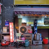 A small street-side setup includes various items such as an air conditioning unit, colorful gas canisters, a few stacked chairs, and various cooking implements like large pots and bowls. There's a neon sign displaying Chinese characters, a partial awning above, and cluttered boxes in the background.