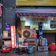 A small street-side setup includes various items such as an air conditioning unit, colorful gas canisters, a few stacked chairs, and various cooking implements like large pots and bowls. There's a neon sign displaying Chinese characters, a partial awning above, and cluttered boxes in the background.