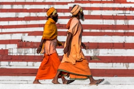 Two men dressed in traditional orange robes and turbans walk side by side in front of a backdrop of red and white striped stairs.