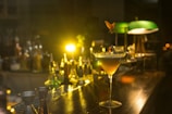 A sophisticated cocktail glass sits on a wooden bar counter, surrounded by various bottles and bar tools. Soft, warm lighting creates a cozy, intimate ambiance, highlighting the yellow and amber hues of the drink. A fruit garnish elegantly decorates the glass, adding a touch of style. In the background, low lighting casts a gentle glow, illuminating green-shaded lamps and adding depth to the scene.