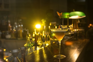 A sophisticated cocktail glass sits on a wooden bar counter, surrounded by various bottles and bar tools. Soft, warm lighting creates a cozy, intimate ambiance, highlighting the yellow and amber hues of the drink. A fruit garnish elegantly decorates the glass, adding a touch of style. In the background, low lighting casts a gentle glow, illuminating green-shaded lamps and adding depth to the scene.