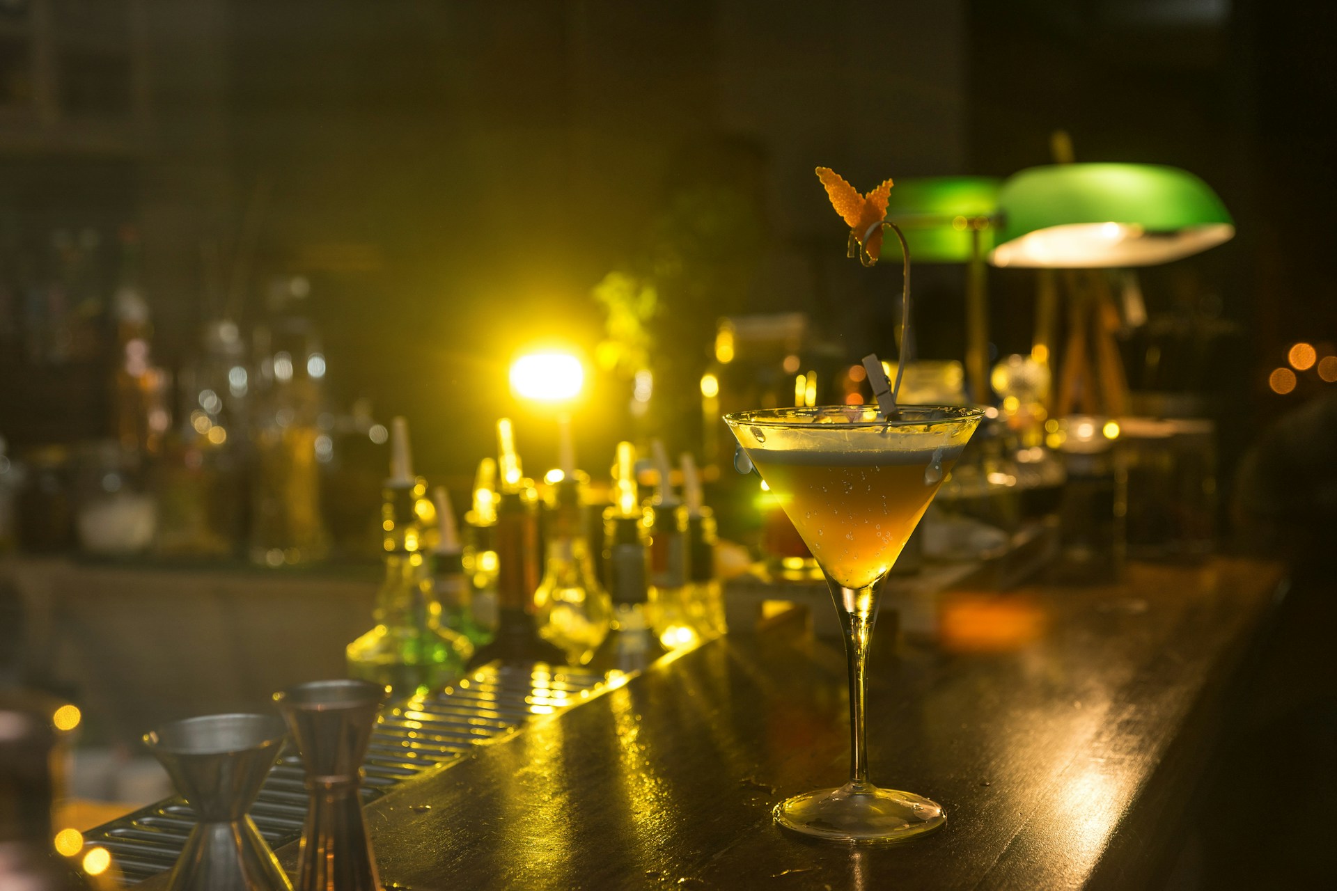 A sophisticated cocktail glass sits on a wooden bar counter, surrounded by various bottles and bar tools. Soft, warm lighting creates a cozy, intimate ambiance, highlighting the yellow and amber hues of the drink. A fruit garnish elegantly decorates the glass, adding a touch of style. In the background, low lighting casts a gentle glow, illuminating green-shaded lamps and adding depth to the scene.