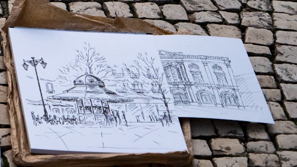 Two pencil sketches are placed on a stone pavement. The sketches depict architectural structures; one features a domed kiosk with nearby street lamps and people, while the other shows a detailed facade of a classical building.