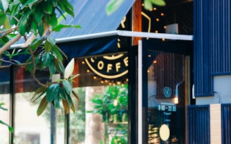 A cozy coffee shop with a modern storefront featuring a large glass window. There is a magnolia tree with large green leaves and a white blossom in the foreground. Warm lights inside the shop create a welcoming atmosphere.