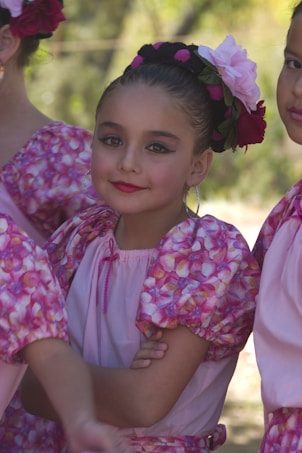 A young girl with her hair styled in a bun adorned with large flowers looks directly at the camera. She wears a dress with a floral pattern in shades of pink, along with matching face makeup. Her expression is composed and confident, and other individuals in similar clothing are visible around her.