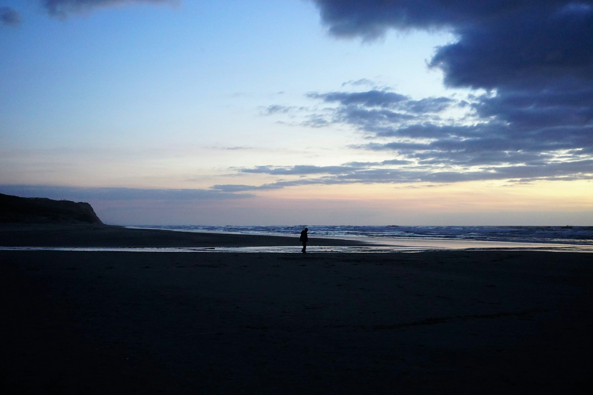 A cinematic frame showing keane austin standing alone on a quiet beach, waves gently rolling in under a twilight sky.