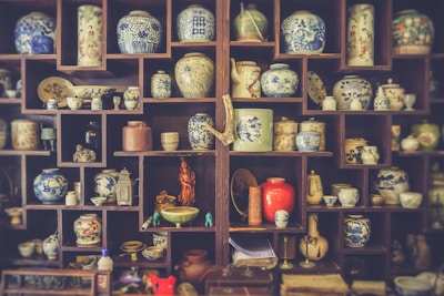A wooden shelf filled with a variety of intricately designed porcelain vases and pots, most displaying blue and white patterns. Some of the shelves contain teapots, small statues, and decorative plates with detailed imagery, showcasing traditional artistry.