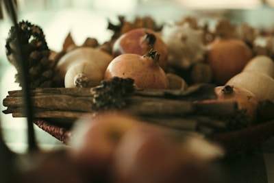 A close-up view of a basket containing various dried fruits and wooden objects. Pomegranates are prominently visible, surrounded by pinecones and other dried botanical elements.