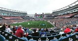 A large sports stadium filled with spectators. The field is green with white markings, and players in red and white uniforms are actively engaged in a game. The stands are packed with people wearing various colored clothing, and some sections of the crowd are more densely populated. Digital screens and advertisements are visible around the stadium, adding to the vibrant atmosphere.