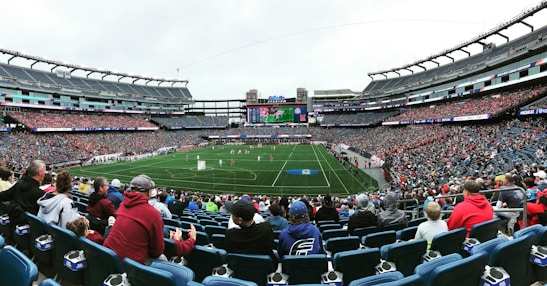 A large sports stadium filled with spectators. The field is green with white markings, and players in red and white uniforms are actively engaged in a game. The stands are packed with people wearing various colored clothing, and some sections of the crowd are more densely populated. Digital screens and advertisements are visible around the stadium, adding to the vibrant atmosphere.