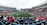 A large sports stadium filled with spectators. The field is green with white markings, and players in red and white uniforms are actively engaged in a game. The stands are packed with people wearing various colored clothing, and some sections of the crowd are more densely populated. Digital screens and advertisements are visible around the stadium, adding to the vibrant atmosphere.