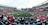 A large sports stadium filled with spectators. The field is green with white markings, and players in red and white uniforms are actively engaged in a game. The stands are packed with people wearing various colored clothing, and some sections of the crowd are more densely populated. Digital screens and advertisements are visible around the stadium, adding to the vibrant atmosphere.