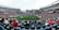 A large sports stadium filled with spectators. The field is green with white markings, and players in red and white uniforms are actively engaged in a game. The stands are packed with people wearing various colored clothing, and some sections of the crowd are more densely populated. Digital screens and advertisements are visible around the stadium, adding to the vibrant atmosphere.