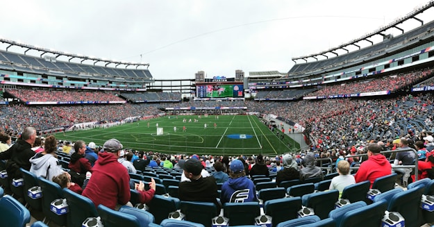 A large sports stadium filled with spectators. The field is green with white markings, and players in red and white uniforms are actively engaged in a game. The stands are packed with people wearing various colored clothing, and some sections of the crowd are more densely populated. Digital screens and advertisements are visible around the stadium, adding to the vibrant atmosphere.