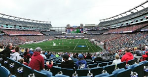 A large sports stadium filled with spectators. The field is green with white markings, and players in red and white uniforms are actively engaged in a game. The stands are packed with people wearing various colored clothing, and some sections of the crowd are more densely populated. Digital screens and advertisements are visible around the stadium, adding to the vibrant atmosphere.