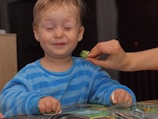 A toddler defiantly pushing away a plate of broccoli with a playful grimace.