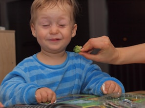 A toddler defiantly pushing away a plate of broccoli with a playful grimace.