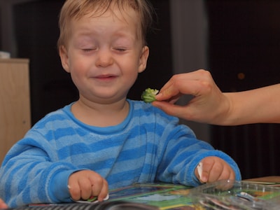 A young child with a joyful expression, wearing a striped blue shirt. A hand is offering a piece of broccoli to the child, who is seated at a table with a toy or book in front of them.