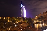 A modern, sail-shaped skyscraper illuminated in vibrant purple and blue lights stands prominently against a night sky. Surrounding the tower, traditional Middle Eastern architecture and lush palm trees are visible, with soft warm lighting reflecting on a body of water. The setting combines modernity with traditional elements, creating a striking and picturesque scene.