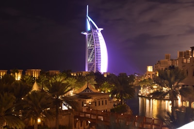 A modern, sail-shaped skyscraper illuminated in vibrant purple and blue lights stands prominently against a night sky. Surrounding the tower, traditional Middle Eastern architecture and lush palm trees are visible, with soft warm lighting reflecting on a body of water. The setting combines modernity with traditional elements, creating a striking and picturesque scene.