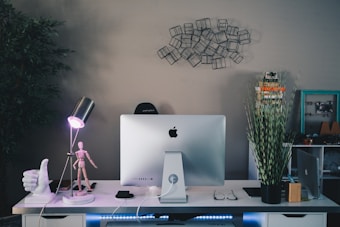 A modern office workspace featuring a large Apple computer on a white desk. Surrounding the computer are various objects, including a stack of books, a potted plant with tall green leaves, a decorative hand sculpture, an adjustable desk lamp, and a wooden mannequin model. A wireframe art piece decorates the wall above a black chair.